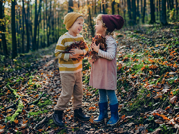 Zwei Kinder stehen in einem herbstlichen Wald. Beide haben trockene Blätter in den Armen.